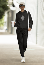Load image into Gallery viewer, Woman wearing a black hoodie and pants with est. 1975 logo, holding a coffee cup and a white bag, walking outdoors.